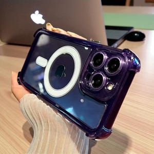 May include: A clear phone case with a purple bumper and a white circular MagSafe charger. The case has a transparent back, showcasing the phone's design. The case has a protective bumper around the edges.