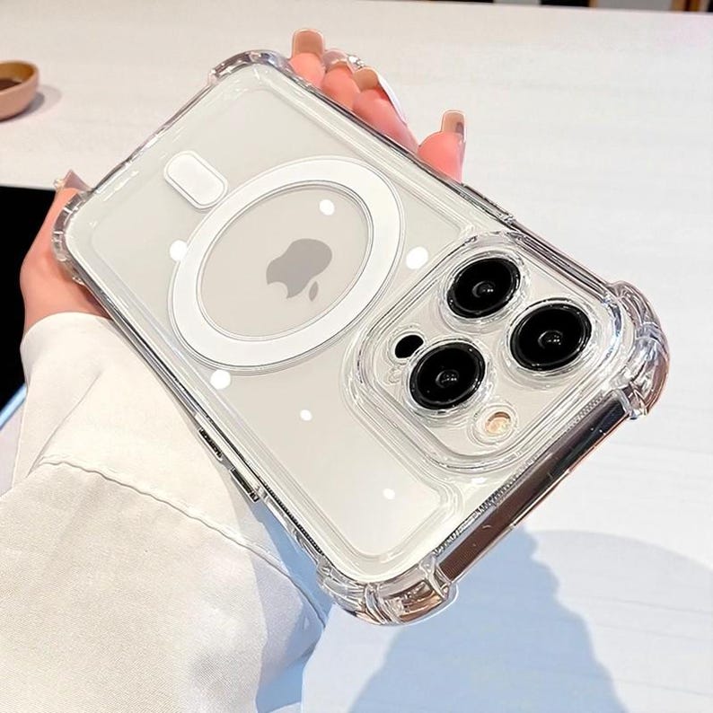 May include: Clear phone case with a protective bumper. The case features a white MagSafe ring and a clear back, showcasing the phone's design. The case has a raised edge around the camera lenses.