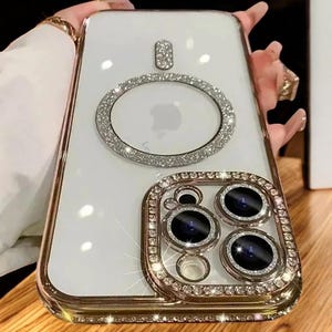 May include: A white phone case with a gold trim and a diamond-encrusted ring around the camera lens. The case has a clear back and a diamond-encrusted ring around the Apple logo.
