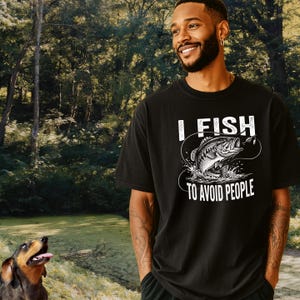 May include: A black short-sleeved t-shirt featuring a white graphic of a fish jumping out of water with a fishing line and hook. The text "I FISH TO AVOID PEOPLE" is printed in bold white capital letters above and below the fish illustration.