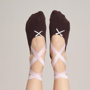 Może przedstawiać: Para brązowych skarpet zaprojektowanych na wzór baletek. Skarpetki mają małą białą kokardkę na palcach i są zabezpieczone jasnoróżowymi wstążkami, które owijają kostki i stopy. Tło jest jednolite, jasnoszare.
