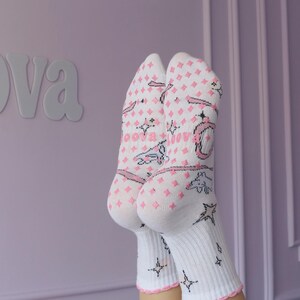 May include: White socks with pink diamond pattern, stars, and text. The socks have a pink scalloped trim. The word "oova" is visible on the socks. The background is a light purple wall.