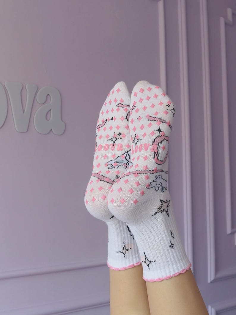May include: White socks with pink diamond pattern, stars, and text. The socks have a pink scalloped trim. The word "oova" is visible on the socks. The background is a light purple wall.
