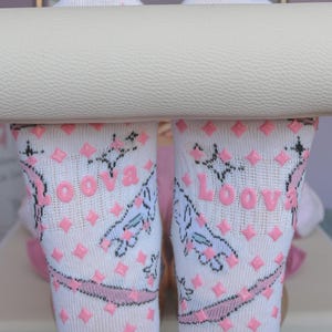 May include: White socks with pink diamond-shaped grips and the word "Loova" printed in pink. The socks feature a pink band and a whimsical design. The socks are designed for comfort and grip.