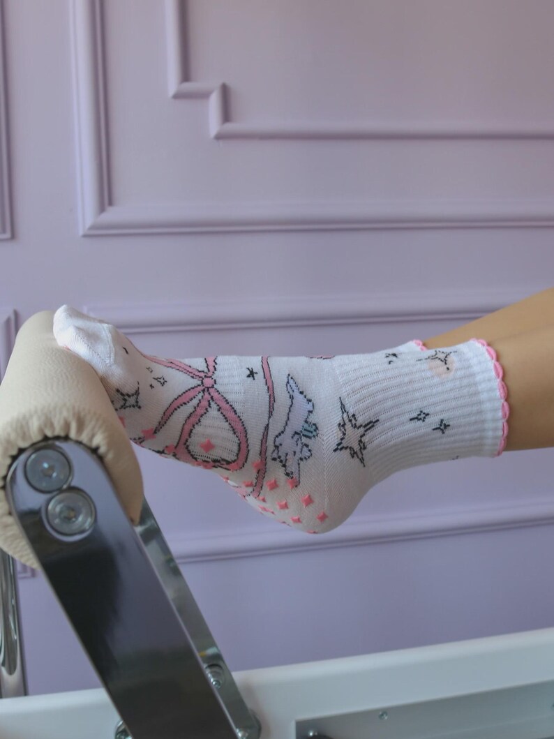 May include: White socks with a pink bow, stars, and cartoon character design. The socks have a pink scalloped trim and pink non-slip grips on the sole. The socks are on a light-colored surface against a lavender wall.