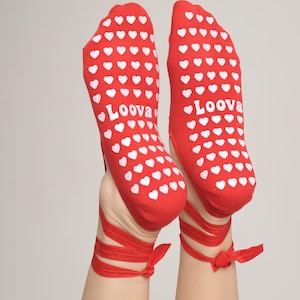 Może przedstawiać: Czerwone skarpetki z białymi sercami i napisem "Loova" na podeszwach. Czerwone wstążki zawiązane wokół kostek. Radosny i stylowy dodatek.