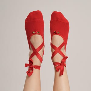 Może przedstawiać: Para czerwonych skarpetek zaprojektowanych tak, aby przypominały baletki. Skarpetki mają mały detal w kształcie róży i są zawiązane czerwoną wstążką wokół kostek. Obraz przedstawia stopy i kostki na gładkim tle.