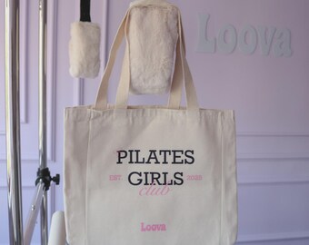 Puede incluir: Un bolso tote de color crema con el texto "PILATES GIRLS CLUB" en negro y rosa. El bolso cuelga frente a una pared lila con la palabra "Loova" en blanco. El bolso tiene correas largas y es parte de un estudio de Pilates.