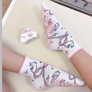 May include: White socks with a pink scalloped trim and a repeating pattern of pink bows, bunnies, and floral accents. A white bunny-shaped pin with a pink bow is also present.