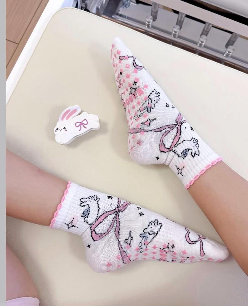 May include: White socks with a pink scalloped trim and a repeating pattern of pink bows, bunnies, and floral accents. A white bunny-shaped pin with a pink bow is also present.