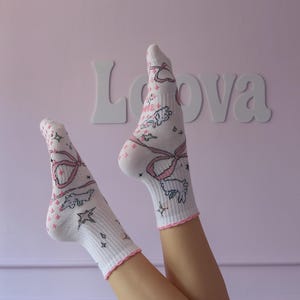 May include: White socks with pink bows, stars, and cartoon animal designs. The socks have pink trim and are against a pink background with the word "Lova" in white letters.