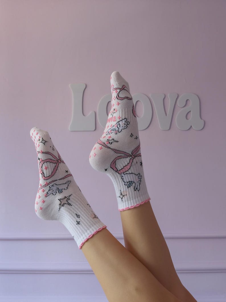 May include: White socks with pink bows, stars, and cartoon animal designs. The socks have pink trim and are against a pink background with the word "Lova" in white letters.