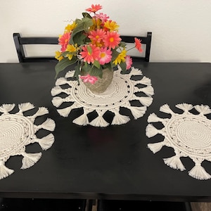 May include: A black dining table with three cream-colored, circular macrame placemats with fringe. A vase of pink, yellow, and orange flowers sits on the center placemat. The table is set against a white wall.