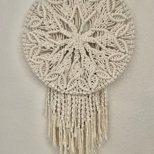 May include: Cream-colored macrame wall hanging. The circular design features intricate knotting patterns, with a central starburst motif and a fringe at the bottom. The macrame is mounted on a metal ring.