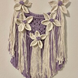 May include: A handmade macrame wall hanging featuring a lavender and cream color scheme. The piece is adorned with five cream-colored, three-dimensional flower accents with lavender centers. The design includes long fringe and a woven top.