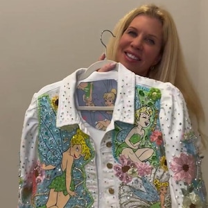 May include: A white denim jacket featuring multiple illustrations of Tinker Bell, embellished with colorful floral appliques, sparkling rhinestones, and sequins. The jacket has a collared neckline, gold-toned buttons, and a patterned inner lining.
