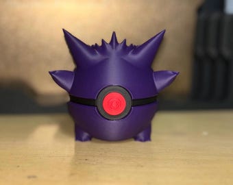May include: A 3D-printed purple figure with a red and black circular design on its front. The figure has pointed ears and small arms. The object is a stylized representation of a cartoon character.