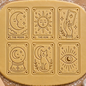 Peut inclure: Une feuille de pâte à biscuits beige clair imprimée de six motifs rectangulaires aux thèmes célestes et mystiques. Les motifs incluent un croissant de lune marqué THE MOON, un soleil marqué THE SUN, une main tenant une carte, une boule de cristal, un chat et un œil.