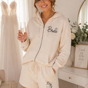 May include: Cream-colored bridal lounge set featuring a zip-up hoodie with "Bride" script and matching shorts with "Mrs. Smith" script. The set is made of a soft material and is perfect for wedding day preparations.