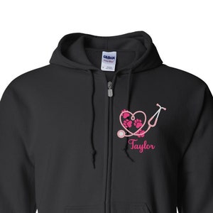 May include: Black zip-up hoodie with a pink heart and stethoscope design. The heart has paw prints inside and the text "Taylor" is below the design.