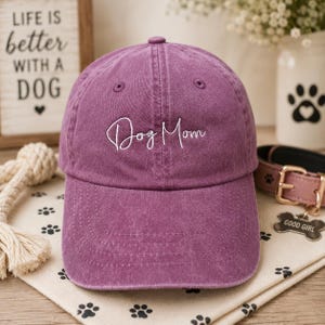 May include: A purple baseball cap with the embroidered text "Dog Mom" in white. The cap is made of a soft, washed fabric. A dog collar and a sign that says "Life is better with a dog" are in the background.