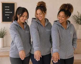 May include: Three people wearing gray zip-up hoodies with personalized embroidery. The hoodies have the names "Amanda", "Melinda", and "KC" embroidered on the chest, and the word "Bridesmaid" on the sleeve. A sign in the background reads "Coffee Scrubs Compassion Repeat".