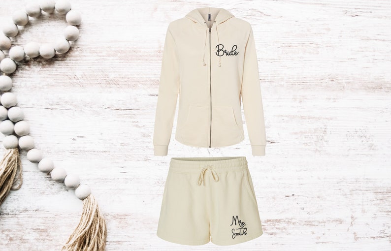 May include: Cream-colored matching loungewear set. The set includes a zip-up hoodie with "Bride" in script and drawstring shorts with "Mrs. Smith" in script. The outfit is displayed on a white wood background.