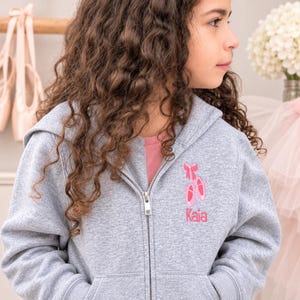 Op de afbeelding: Een grijs gemêleerde zip-up hoodie met een geborduurd ontwerp op de linkerborst. Het borduursel toont een paar roze balletschoenen met een bijpassende roze strik erboven en de naam Kaia in roze letters eronder gestikt.