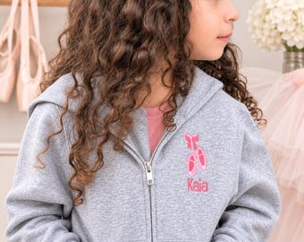 May include: A heather gray zip-up hooded sweatshirt featuring an embroidered design on the left chest. The embroidery depicts a pair of pink ballet slippers with a matching pink bow above them and the name Kaia stitched in pink lettering below.