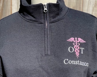 May include: Black quarter-zip sweatshirt with a pink embroidered medical symbol, the letters "OT" and the name "Constance" in white.