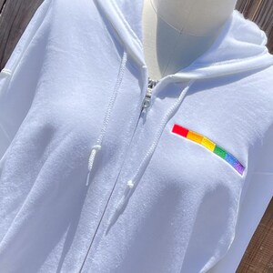 May include: White zippered hoodie with a rainbow embroidered patch on the front.