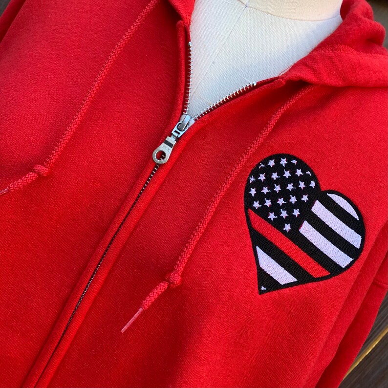 K&ouml;nnte beinhalten: Roter Kapuzenpullover mit Rei&szlig;verschluss und einem herzf&ouml;rmigen Patch mit einer amerikanischen Flagge mit einem roten Streifen in der Mitte.