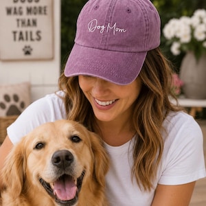 May include: A dusty rose baseball cap with the script "Dog Mom" embroidered on the front. The cap is worn by a person smiling, holding a golden retriever. A sign in the background reads "Dogs wag more than tails."