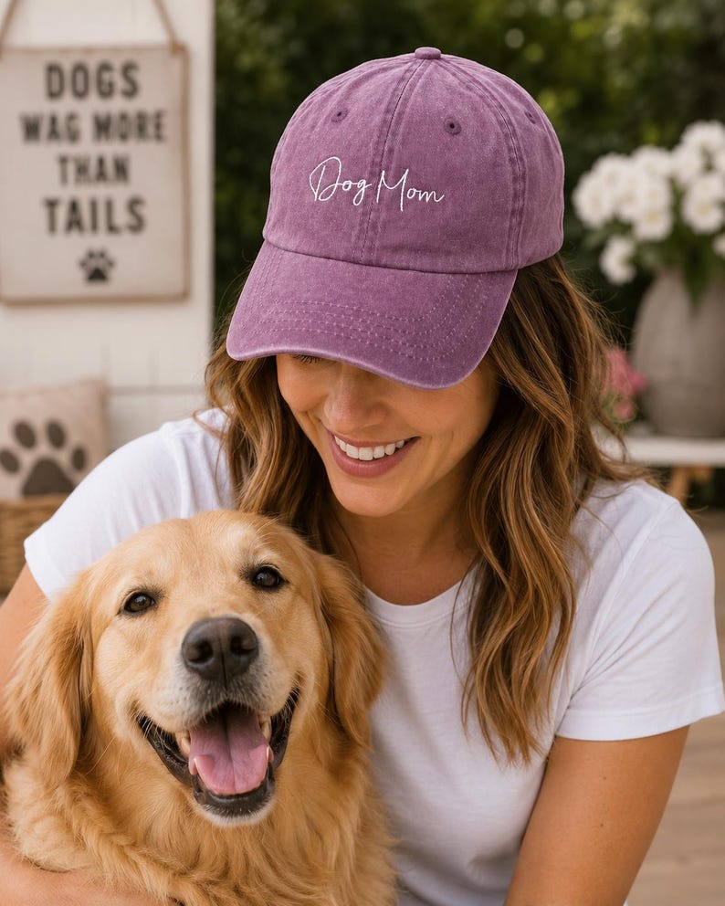 May include: A dusty rose baseball cap with the script "Dog Mom" embroidered on the front. The cap is worn by a person smiling, holding a golden retriever. A sign in the background reads "Dogs wag more than tails."
