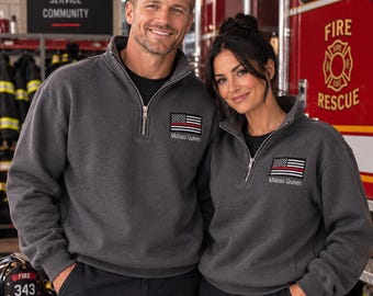 May include: Two charcoal gray quarter-zip sweatshirts featuring an embroidered American flag patch with a thin red line and the text Mabas Queen underneath. The sweatshirts have a silver-toned metal zipper at the collar and long sleeves.
