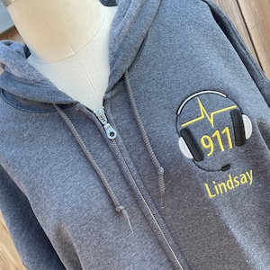 May include: A gray zippered hoodie with a yellow embroidered design of a headset with a heartbeat and the number 911. The name Lindsay is also embroidered in yellow below the headset.