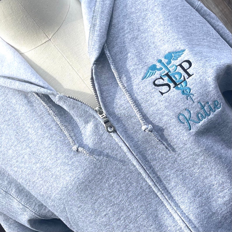 Op de afbeelding: Grijze hoodie met rits, een geborduurd medisch symbool in blauw en zwart en de tekst "SLP Katie".