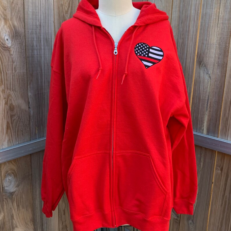 K&ouml;nnte beinhalten: Roter Kapuzenpullover mit Rei&szlig;verschluss und einem Herz-Design in den Farben der amerikanischen Flagge in Schwarz und Wei&szlig;.