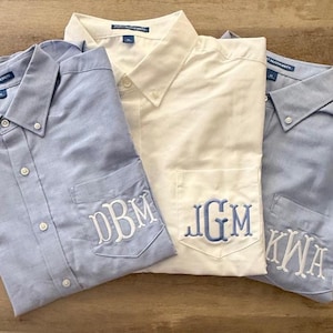 Puede incluir: Tres camisas abotonadas en azul claro y crema. Cada camisa tiene un bolsillo con monograma con iniciales bordadas en blanco o azul. Las camisas están dobladas y dispuestas sobre una superficie de madera.