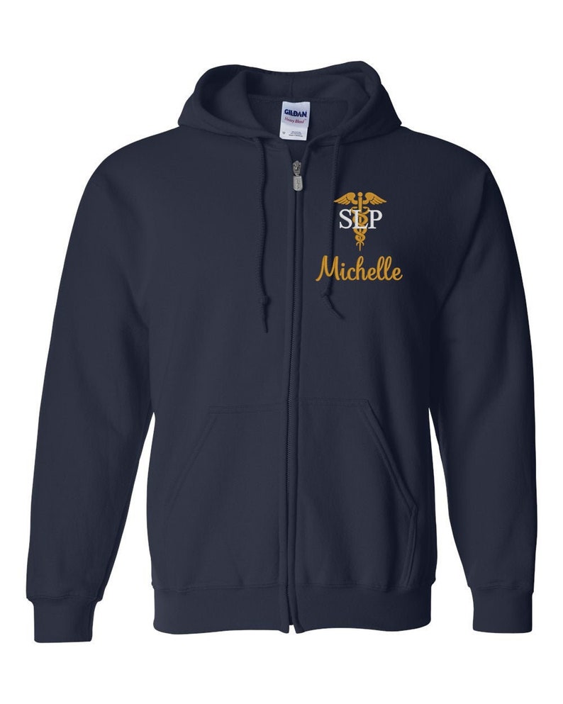 Op de afbeelding: Donkerblauwe hoodie met rits en een geborduurd medisch symbool in goud, de letters "SLP" en de naam "Michelle".