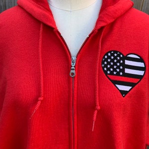 K&ouml;nnte beinhalten: Roter Rei&szlig;verschluss-Hoodie mit einem herzf&ouml;rmigen amerikanischen Flaggen-Patch mit einem d&uuml;nnen roten Streifen in der Mitte.