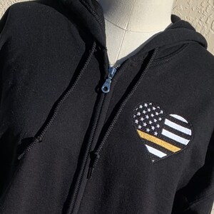 May include: Black zippered hoodie with a heart-shaped embroidered patch featuring a thin yellow line over an American flag.