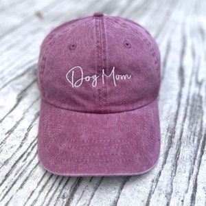 May include: A dusty rose baseball cap with the embroidered text "Dog Mom" in white script. The cap has a curved brim and a classic six-panel design. The hat is made of a soft, textured fabric.