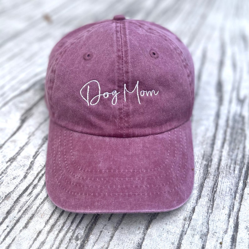 May include: A dusty rose baseball cap with the embroidered text "Dog Mom" in white script. The cap has a curved brim and a classic six-panel design. The hat is made of a soft, textured fabric.
