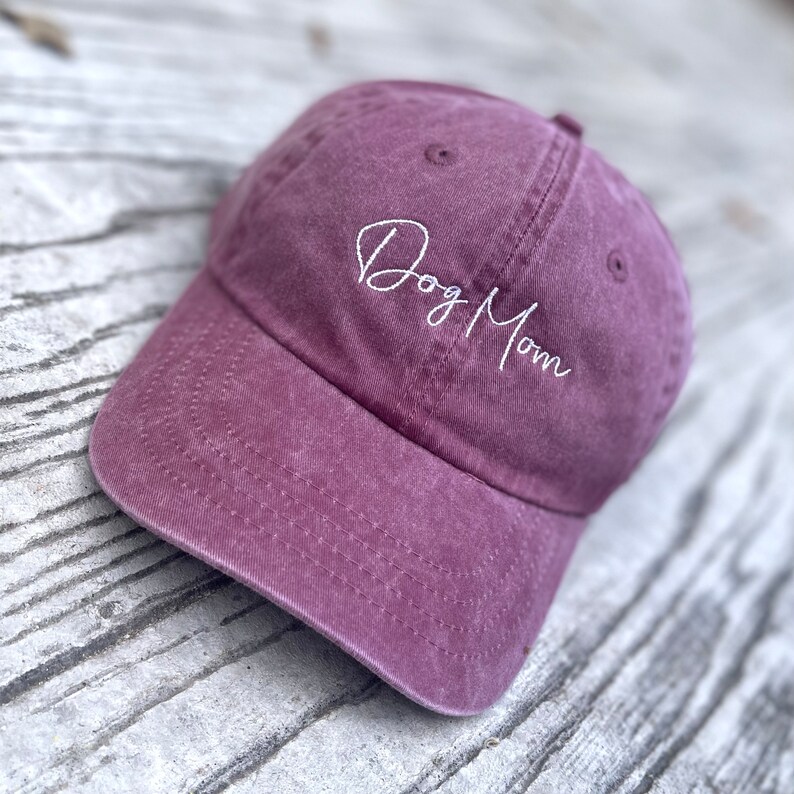 May include: A dusty rose baseball cap with the embroidered text "Dog Mom" in white script. The cap has a curved brim and a fabric strap closure on the back. The hat is made of a soft, worn material.