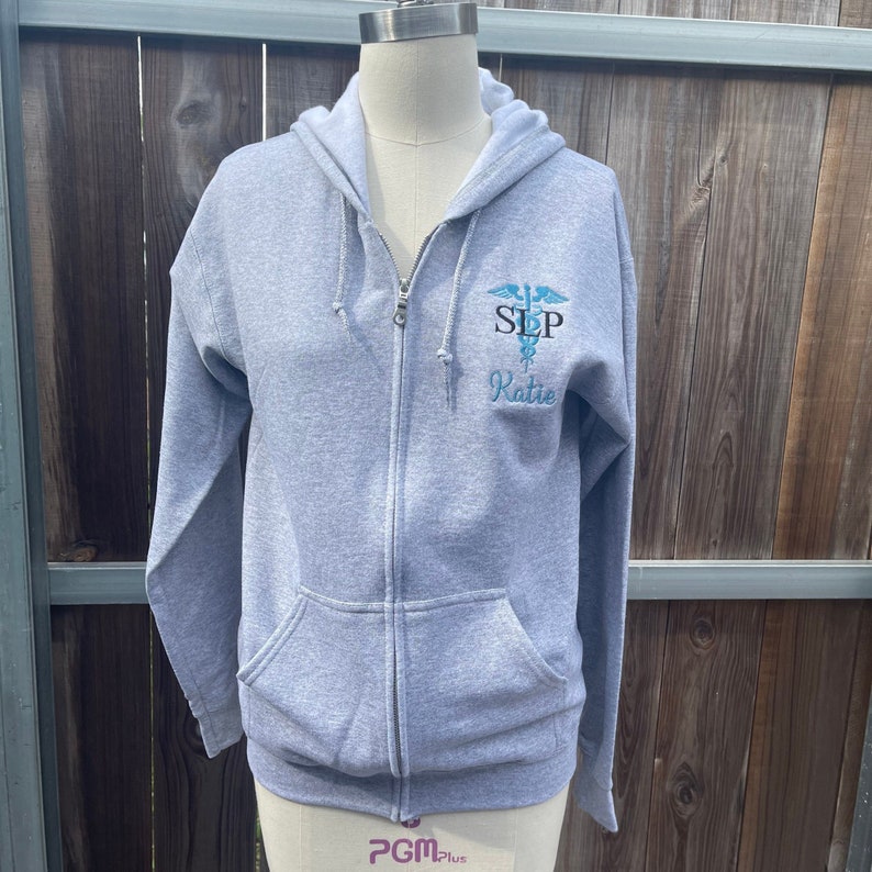 Op de afbeelding: Een grijze hoodie met rits met een blauw en zwart geborduurd medisch symbool en de tekst "SLP Katie".