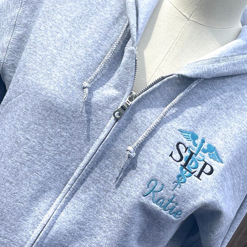 Op de afbeelding: Grijze hoodie met rits, een geborduurd medisch symbool in blauw en zwart en de tekst "SLP Katie".