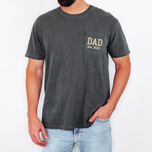 May include: Dark gray t-shirt with a pocket on the chest. The pocket features the word "DAD" above "Est. 2025" in a gold color. The shirt has short sleeves and a crew neck.