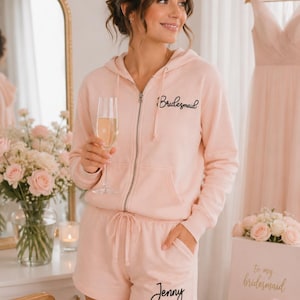 May include: A light pink zip-up hoodie and matching shorts set. The word "Bridesmaid" is scripted on the chest of the hoodie, and the name "Jenny" is scripted on the shorts. The set is made of a soft material and is part of a bridal party theme.