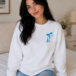Op de afbeelding: Wit sweatshirt met een blauwe strik en de letter "J" op de voorkant. Het sweatshirt heeft een ronde hals en wordt gecombineerd met een spijkerbroek. De foto is binnenshuis genomen.